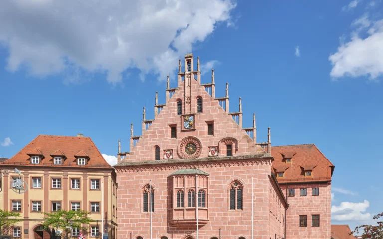 Gotisches Rathaus in Sulzbach-Rosenberg, ein historisches Gebäude mit Steinfassade, Treppengiebel, Uhr und Wappen, unter blauem Himmel mit Wolken.