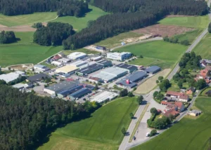 Luftaufnahme zeigt Gewerbegebiet mit mehreren Produktionshallen unterschiedlicher Größe, Parkplätze mit Fahrzeugen, umgeben von dichten Waldgebieten, grünen Wiesen und einzelnen Häusern