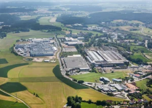 Luftaufnahme zeigt Sulzbach-Rosenberg mit mehreren großen Produktionshallen, Parkplätzen, umgeben von grünen Wiesen, Waldgebieten und landwirtschaftlichen Feldern in hügeligem Terrain
