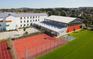 Moderne Pestalozzi Grundschule in Sulzbach-Rosenberg mit weißem Hauptgebäude, roter Sporthalle und Sportplatz, umgeben von grüner Landschaft